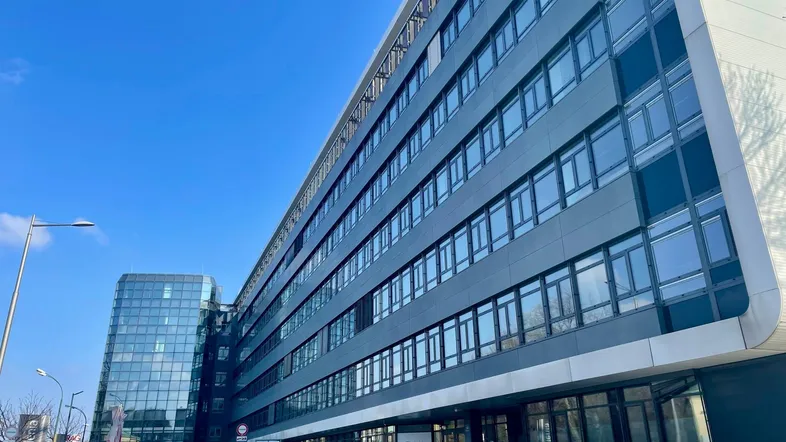 Modernes Bürogebäude mit Glasfassade und davorliegendem Parkplatz unter blauem Himmel.