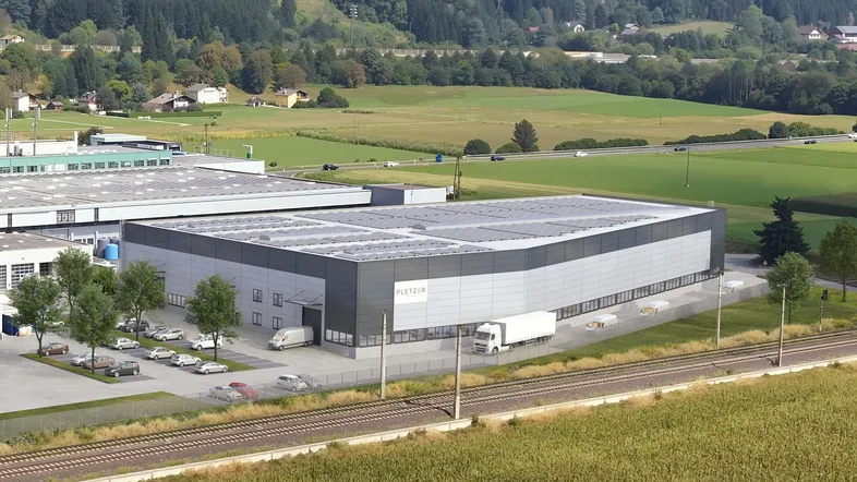 Luftaufnahme eines modernen Industriegebäudes mit großem Parkplatz und umliegender grüner Landschaft.