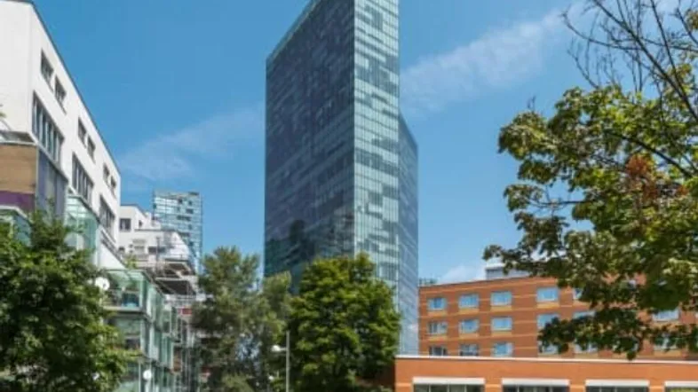 Moderne Hochhausfassade aus Glas in einer städtischen Umgebung mit umliegenden Gebäuden und Bäumen.