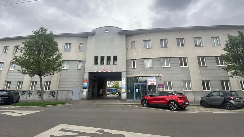 Gebäudeansicht mit mehreren Stockwerken, Durchfahrt und geparkten Autos vor der Fassade unter bewölktem Himmel.