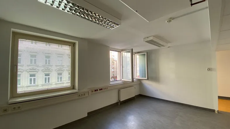 Leerer Büroraum mit mehreren Fenstern, Jalousien und Deckenbeleuchtung. Der Raum hat einen grauen Boden und weiße Wände.