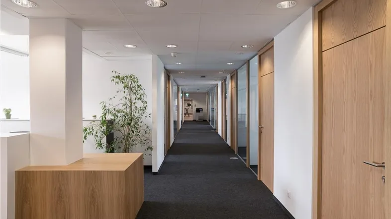 Langer Bürogang mit Teppichboden, Holztüren und Glaswänden, der zu verschiedenen Büros führt.
