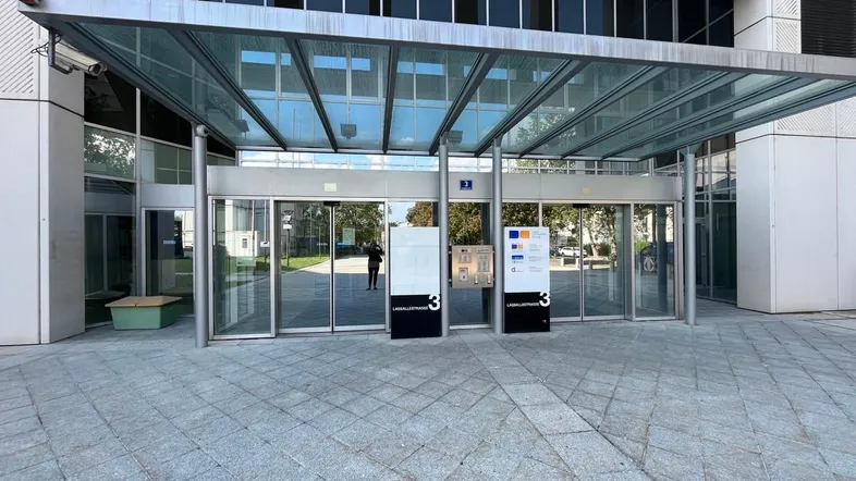 Moderner Gebäudeeingang mit Glasfassade, automatischen Türen und Überdachung, umgeben von Pflastersteinen.