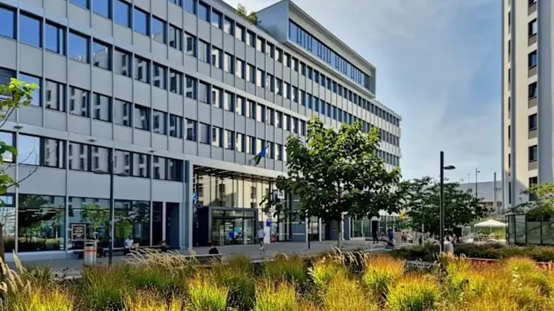 Modernes Bürogebäude mit großen Fenstern und begrüntem Vorplatz unter blauem Himmel.