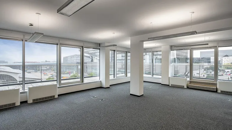 Geräumiges, leeres Büro mit Teppichboden und großen Fenstern, die viel Tageslicht hereinlassen.