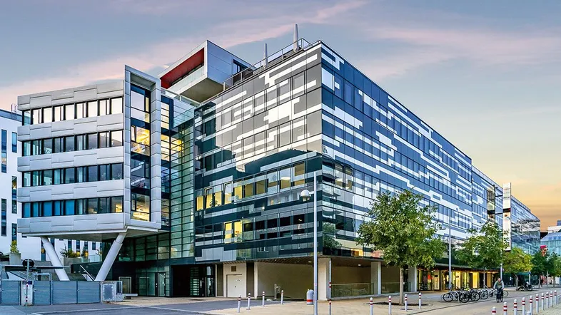 Modernes Bürogebäude mit Glasfassade und markanter Architektur bei Sonnenuntergang, umgeben von Bäumen.