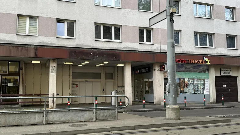 Fassade eines Mehrfamilienhauses mit Geschäften im Erdgeschoss und Straßenbahnschienen im Vordergrund einer städtischen Umgebung.