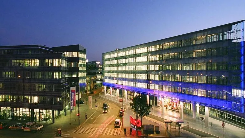 Moderne Bürogebäude mit beleuchteten Fenstern und einer belebten Straße bei Dämmerung.