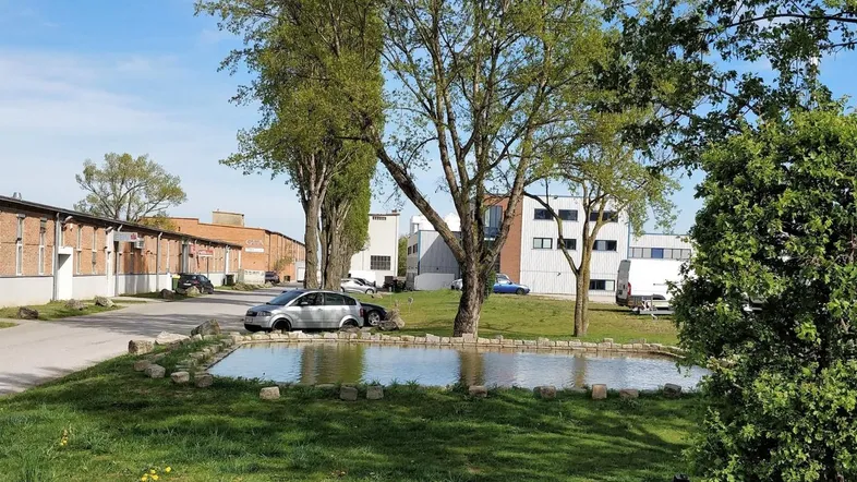 Außenansicht von Industriegebäuden mit einem kleinen Teich und Bäumen in einer grünen Umgebung.