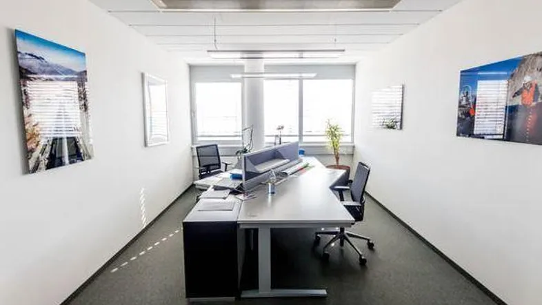 Helles und modernes Büro mit großem Schreibtisch, Teppichboden und natürlichem Lichteinfall durch Fenster.