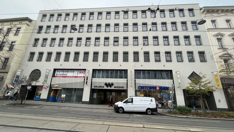 Weißes Geschäftsgebäude mit mehreren Etagen, großen Fenstern und Ladenlokalen im Erdgeschoss. Ein Lieferwagen parkt davor.