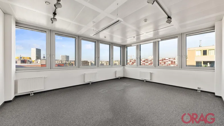 Helles Eckbüro mit Teppichboden und großen Fenstern, die einen weiten Blick über die Stadt bieten.