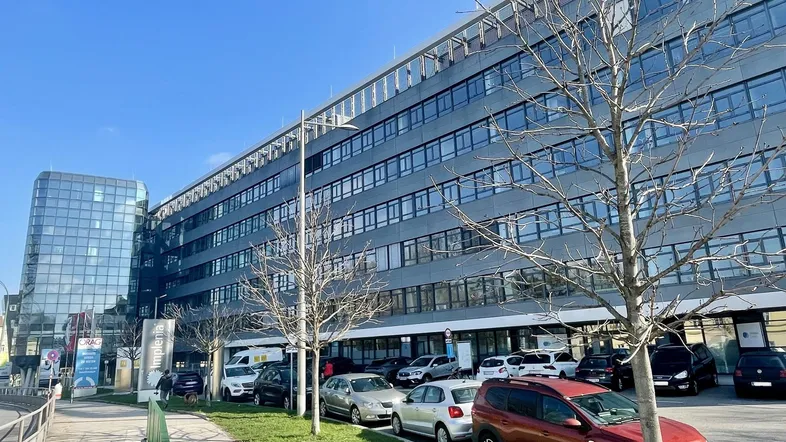 Modernes Bürogebäude mit Glasfassade und Parkplätzen davor unter blauem Himmel.