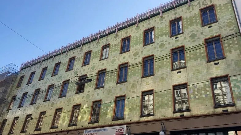Fassade eines Altbaus mit markanten grünen Fliesen und zahlreichen Fenstern in einem städtischen Umfeld.