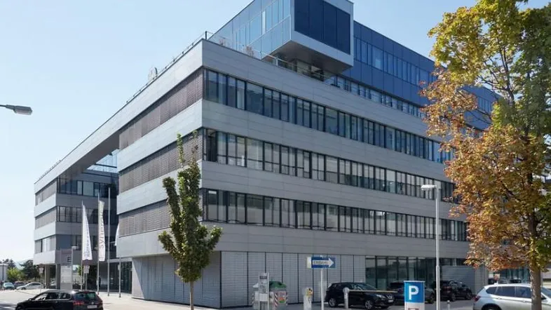 Modernes Bürogebäude mit mehreren Etagen, großen Fenstern und Parkplätzen davor unter blauem Himmel.