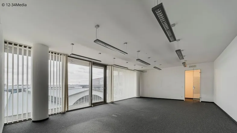Geräumiges, leeres Büro mit großen Fenstern, vertikalen Jalousien und Blick auf die Umgebung.