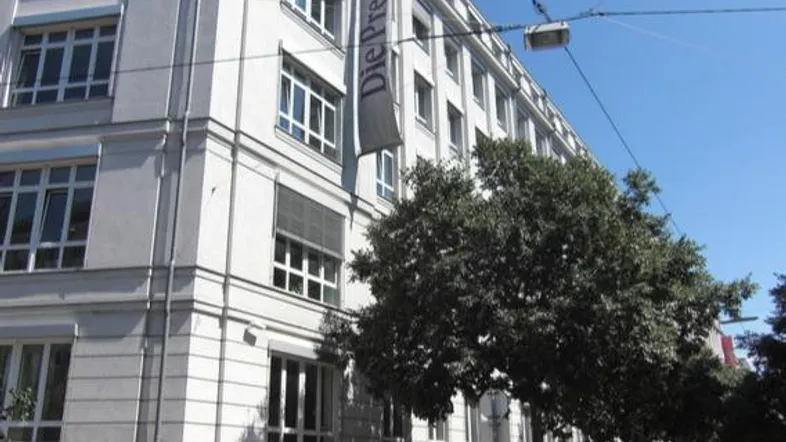 Weißes Gebäude mit vielen Fenstern und einem Banner, umgeben von Bäumen und klarem Himmel in einer städtischen Umgebung.