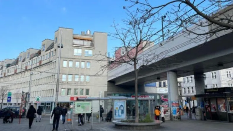 Blick auf die belebte Straße mit Gebäuden, einer Bushaltestelle und einer erhöhten Bahntrasse.
