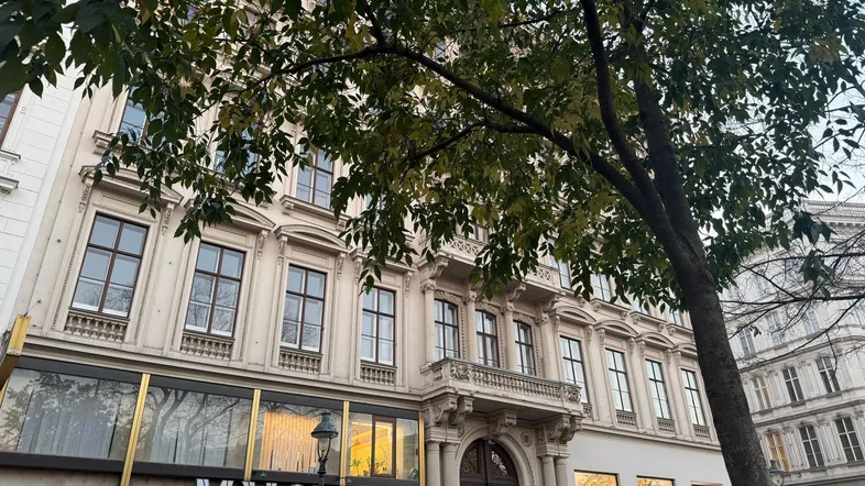 Historisches Gebäude mit klassischer Fassade und großen Fenstern, umgeben von Bäumen und Geschäften.