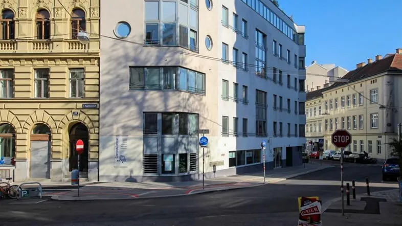 Straßenecke mit einem modernen Gebäude und einem angrenzenden Altbau unter blauem Himmel.