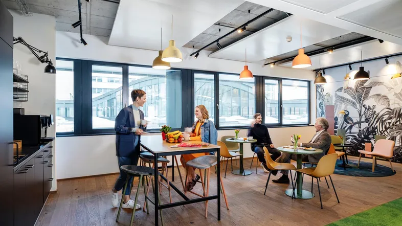 Moderner Büropausenraum mit Holzboden, großen Fenstern und verschiedenen Sitzbereichen für Mitarbeiter.