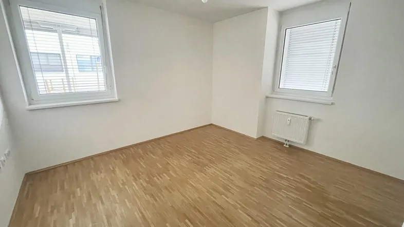 Helles, leeres Zimmer mit Holzparkettboden, weißen Wänden und zwei Fenstern.