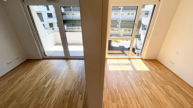 Helles Zimmer mit Holzboden und großen Fenstern, die auf einen Balkon blicken.