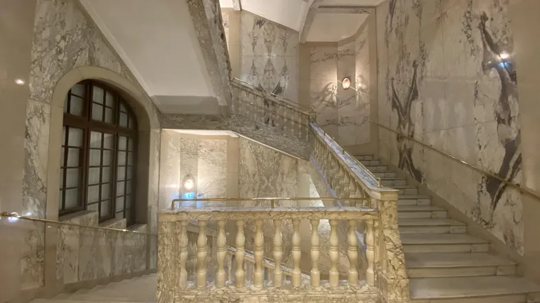 Elegantes Treppenhaus mit Marmorverkleidung, kunstvollen Balustraden und einem großen Bogenfenster, das Tageslicht spendet.