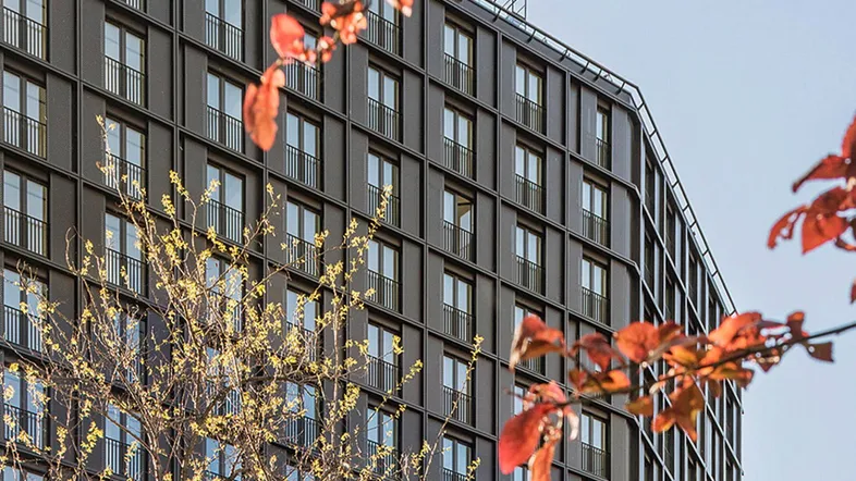 Modernes Gebäude mit dunkler Fassade und zahlreichen Fenstern, umgeben von Bäumen mit Herbstlaub.
