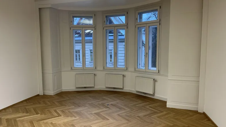 Geräumiger, leerer Raum mit Fischgrätparkett und großen Bogenfenstern, die viel Tageslicht hereinlassen.