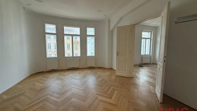 Geräumiges Zimmer mit Fischgrätparkett, hohen Decken und großen Fenstern, die viel Tageslicht hereinlassen.