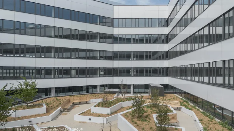 Moderner Innenhof eines Bürogebäudes mit begrünten Beeten und Sitzgelegenheiten, umgeben von großen Fensterfronten.