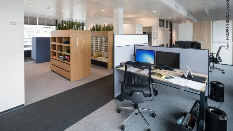 Modernes Großraumbüro mit ergonomischen Arbeitsplätzen, Holzelementen und Pflanzen zur Raumtrennung.