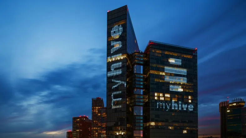 Moderne Hochhausfassade bei Dämmerung mit beleuchteter Schrift und Stadtlichtern im Hintergrund, die eine dynamische Atmosphäre schafft.