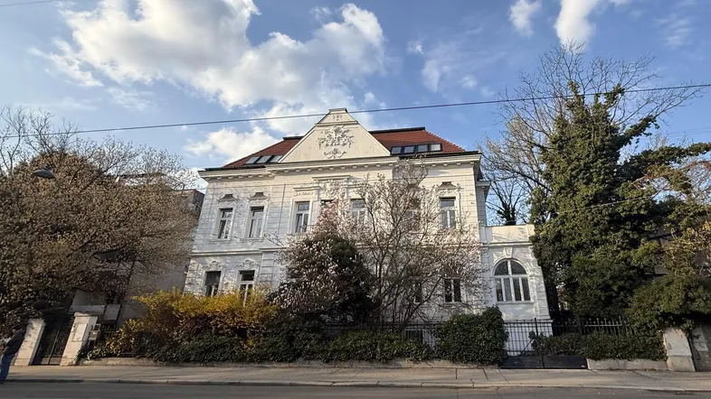 Historisches Gebäude mit klassischer Architektur und Stuckfassade, umgeben von Bäumen und Sträuchern an einer Straße.