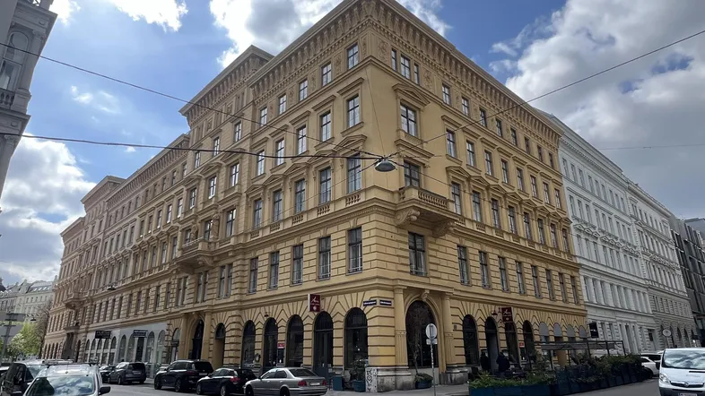 Historisches Gebäude an einer Straßenecke mit klassischer Architektur und vielen Fenstern unter bewölktem Himmel.