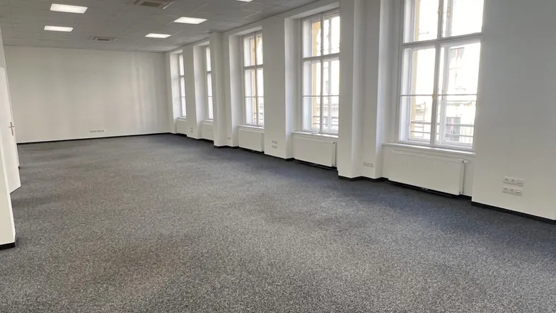 Geräumiger, leerer Büroraum mit Teppichboden, vielen Fenstern und heller Deckenbeleuchtung.