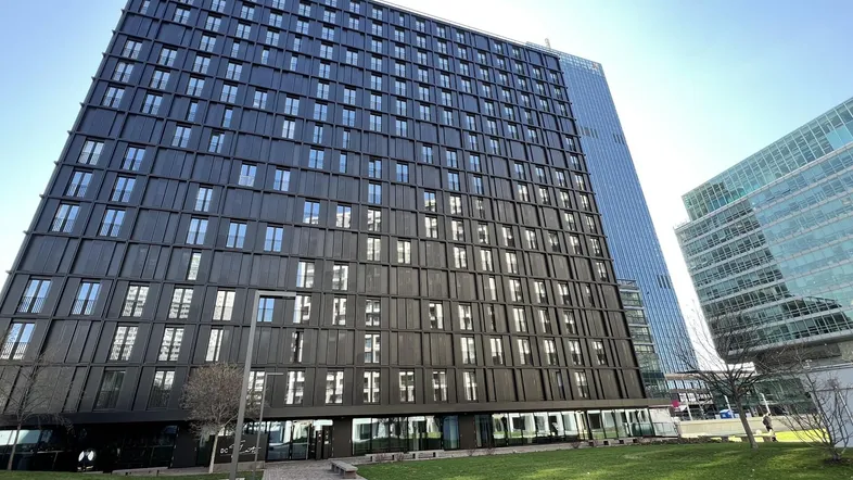 Modernes Hochhaus mit dunkler Fassade und vielen Fenstern, umgeben von Grünflächen und einem Gehweg.