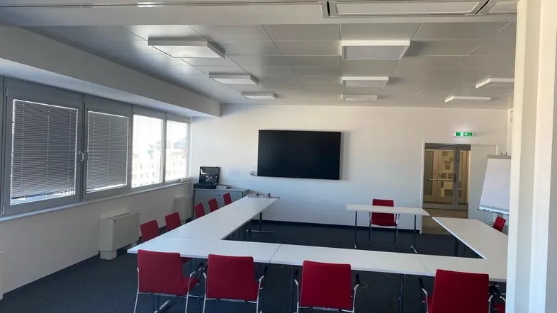 Moderner Konferenzraum mit U-förmiger Tischordnung, großem Bildschirm und natürlichem Lichteinfall durch Fenster.