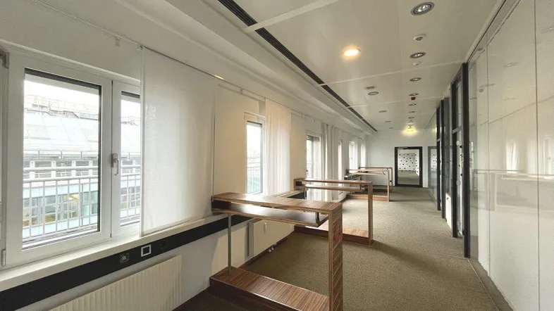 Langer Bürogang mit Teppichboden, großen Fenstern und modernen Holztischen entlang der Wand.