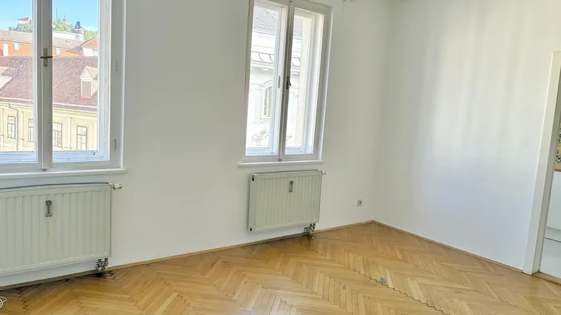 Helles Zimmer mit Fischgrätparkett, zwei großen Fenstern und Blick auf die Umgebung.
