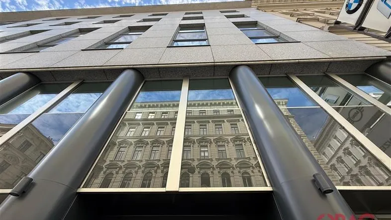 Moderne Bürogebäude-Fassade mit großen Glasflächen, die die gegenüberliegenden historischen Gebäude widerspiegeln.