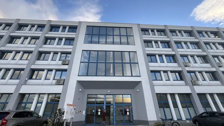 Modernes Bürogebäude mit großer Glasfassade und Eingangsbereich, umgeben von Parkplätzen unter blauem Himmel.