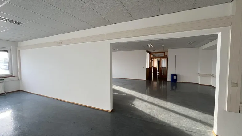 Geräumiges, leeres Büro mit hellen Wänden, abgehängter Decke und dunklem Bodenbelag, durchflutet von natürlichem Licht.