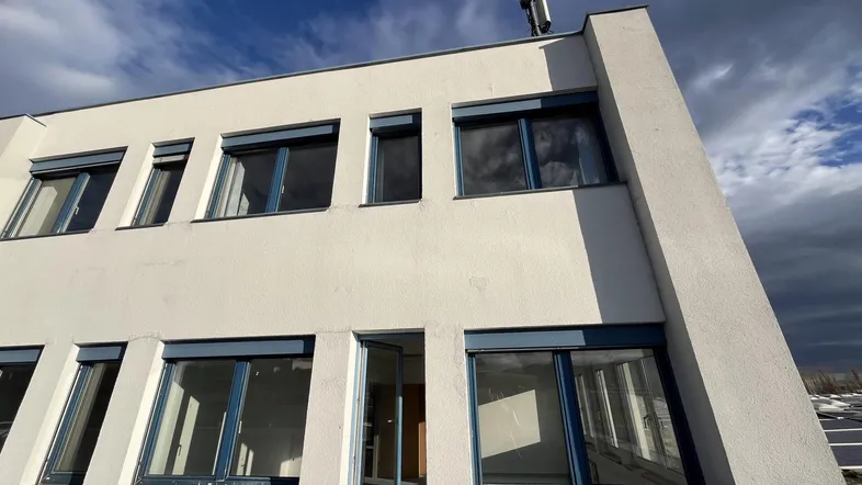 Fassade eines modernen Gebäudes mit blauen Fensterrahmen, Rollläden und einer Antenne auf dem Dach unter bewölktem Himmel.