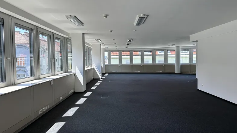 Geräumiges, leeres Büro mit großen Fenstern, Teppichboden und viel Tageslicht.