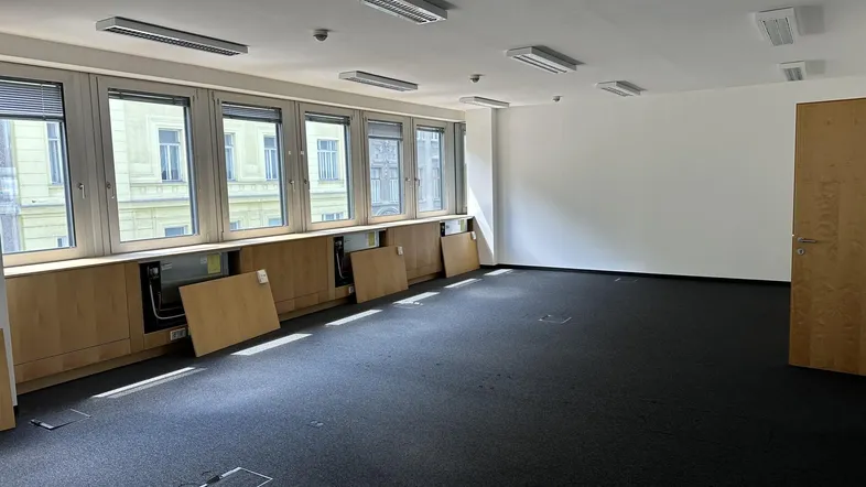 Geräumiges, leeres Büro mit Teppichboden, großen Fenstern und Holzverkleidung unter den Fenstern.