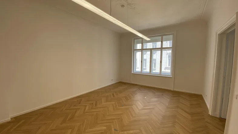 Geräumiges Zimmer mit elegantem Fischgrätparkett, hohen Decken und einem großen Fenster, das viel Tageslicht hereinlässt.