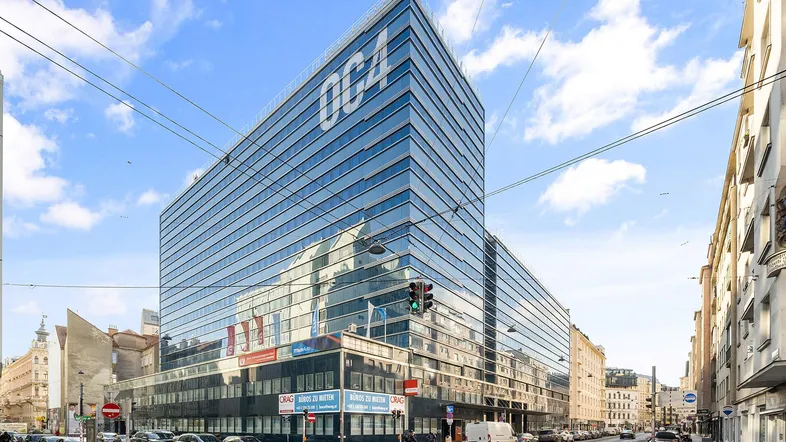 Modernes Bürogebäude mit Glasfassade und blauer Beschriftung an einer belebten Stadtstraße unter blauem Himmel.