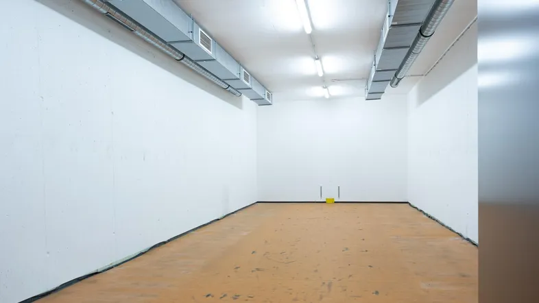 Leerer Gewerberaum mit weißen Wänden, braunem Boden und sichtbaren Lüftungskanälen an der Decke.